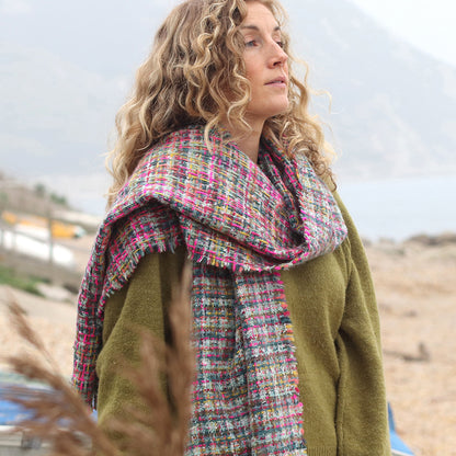 Woman with curly hair, wearing an olive green jumper and a multicoloured woven scarf made from vegan materials, stands outdoors by a body of water with mountains in the background.