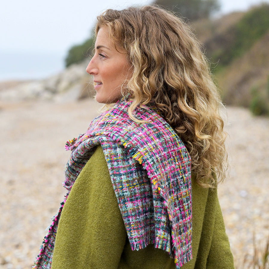 A woman with curly blonde hair, dressed in a green coat and a multicoloured woven scarf made from vegan materials, stands on a rocky beach looking to the left.