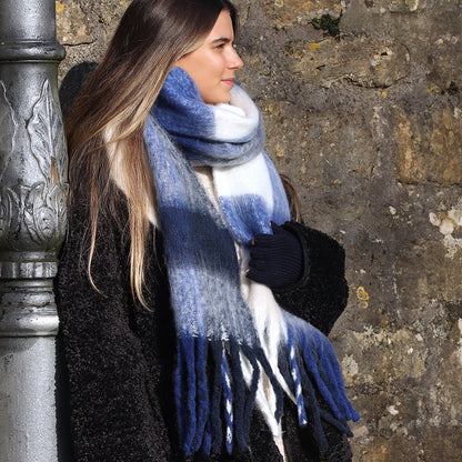 Woman with long hair wearing a cosy winter accessory—a thick blue and white striped scarf with a soft texture—and black coat stands outdoors beside a stone wall.