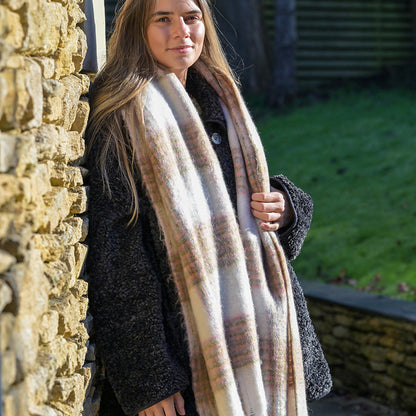 A woman in a dark coat and beige chunky checked scarf stands beside a stone wall outdoors on a sunny day, her cosy accessory adding warmth and charm to her appearance.