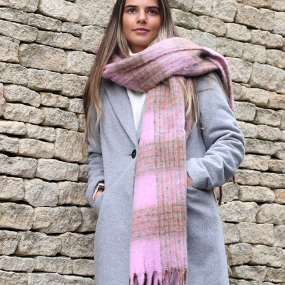 A person in a grey coat and a chunky checked scarf stands before a stone wall, hands tucked into their coat pockets.
