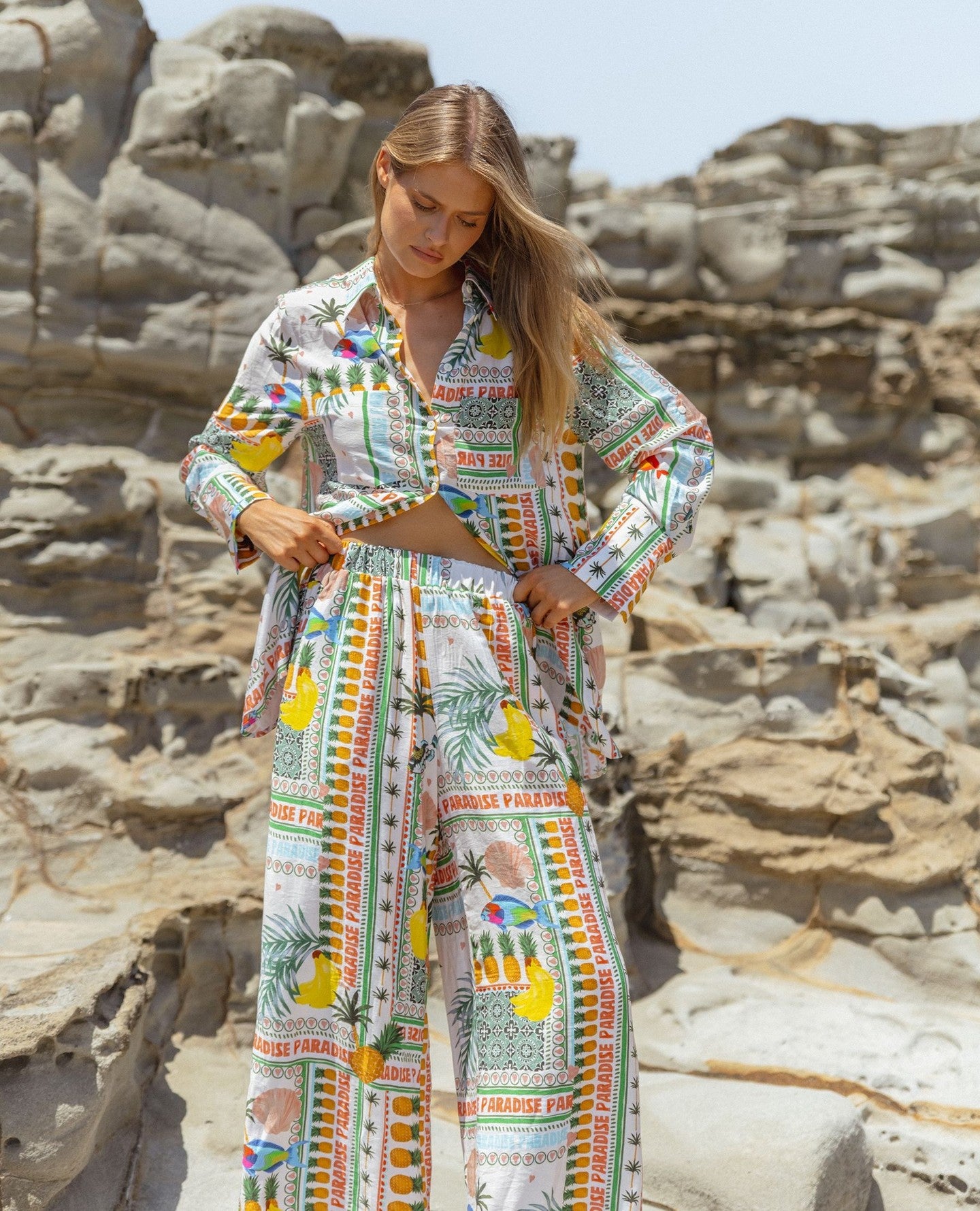 A woman stands on rocky ground wearing a colourful Paradise Print shirt and wide-leg trousers featuring fruit and text designs, highlighting sustainable fashion.