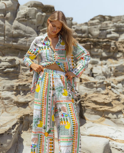 A woman stands on rocky ground wearing a colourful Paradise Print shirt and wide-leg trousers featuring fruit and text designs, highlighting sustainable fashion.