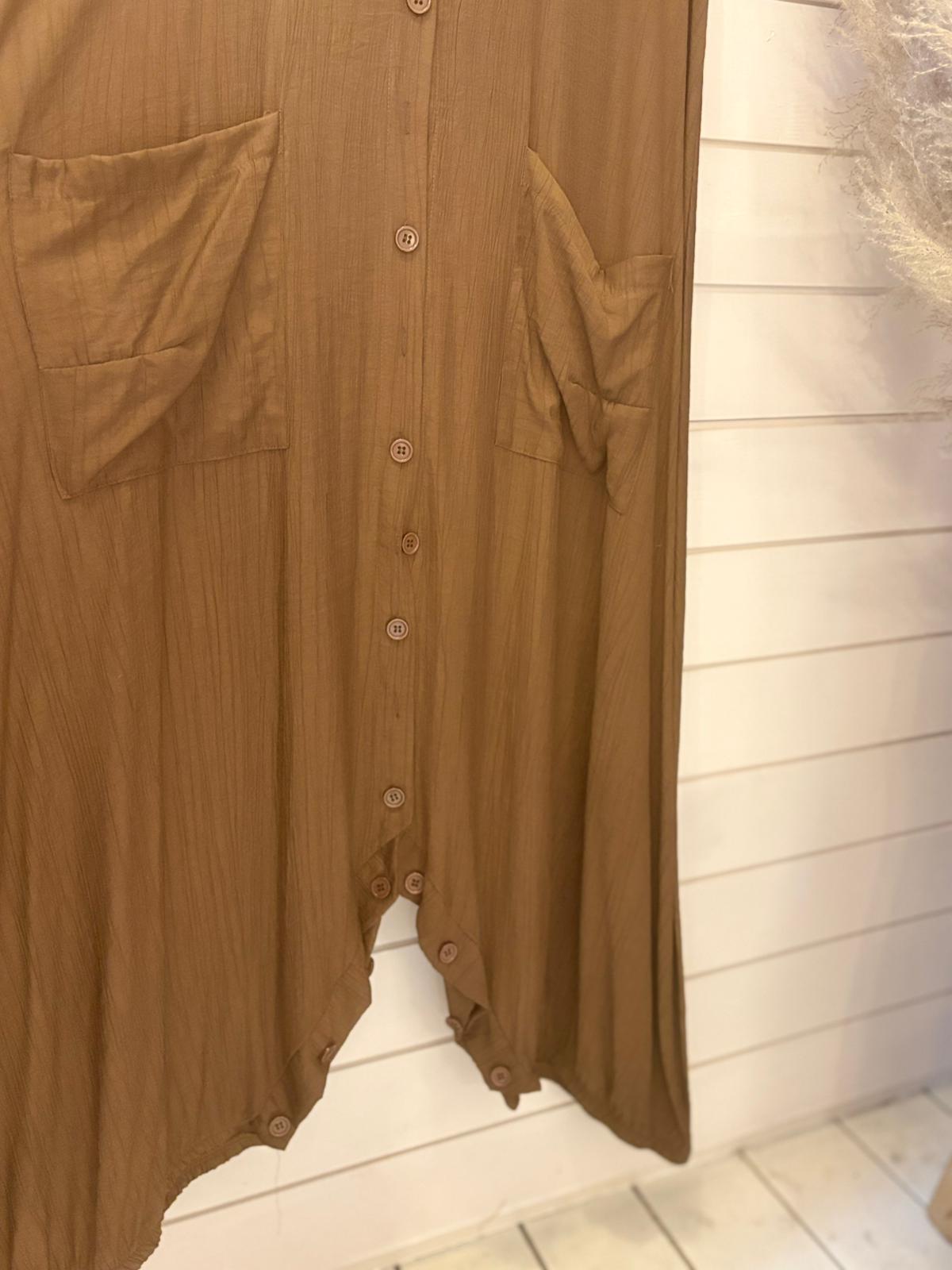 Brown jumpsuit dress with a long silhouette, featuring two large front pockets, hangs against a white wooden wall. Only the lower part of this one-size-fits-all garment is visible.