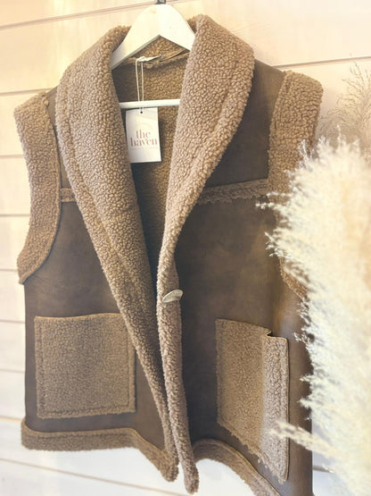 A brown faux wool waistcoat with large front pockets and a shawl collar hangs on a white hanger against a wooden wall.