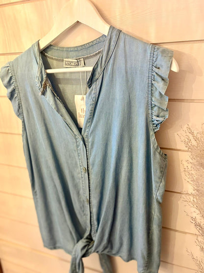 A sleeveless, light blue vegan denim blouse with ruffled shoulders and a front tie, hanging on a white hanger against a wooden wall.