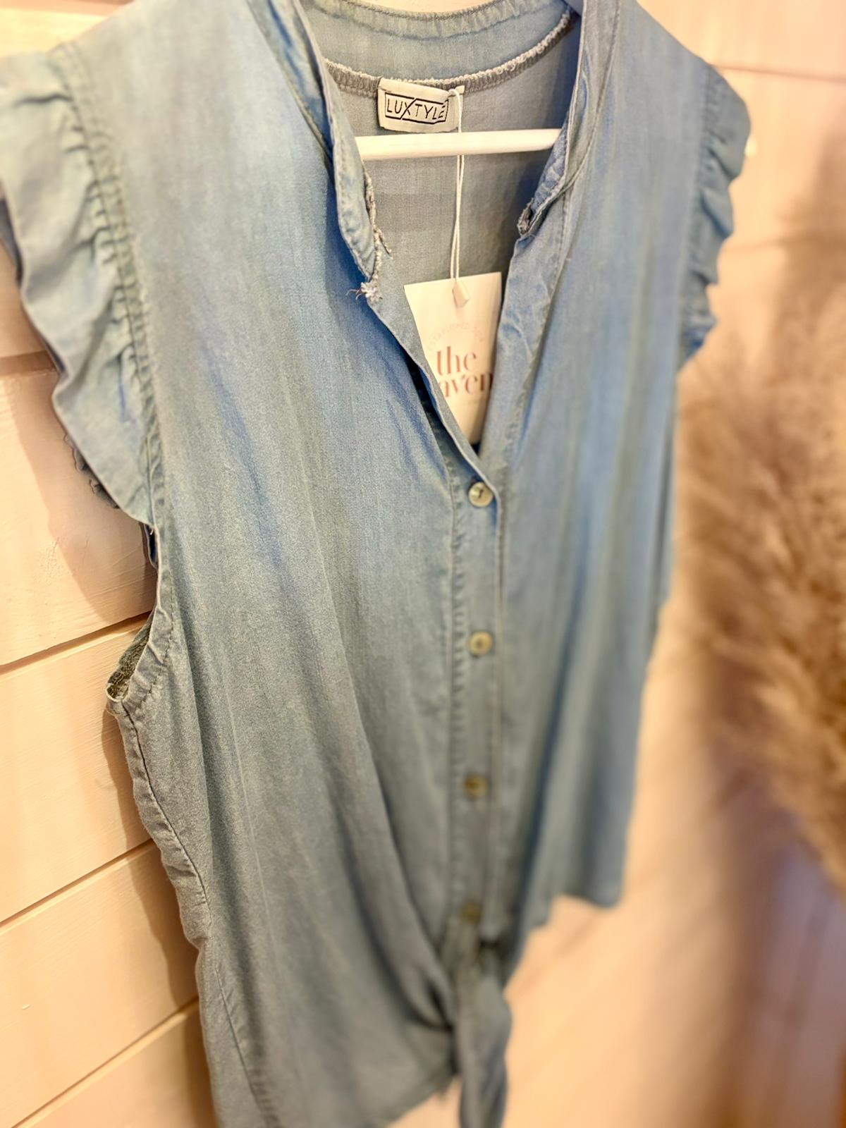 Sleeveless light denim shirt with ruffled armholes and a tied front, made from vegan denim, hanging on a white hanger against a wooden wall.