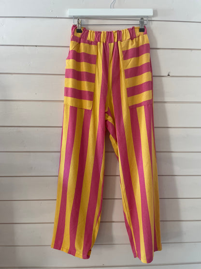Loose-fitting, vegan-friendly trousers with vivid pink and yellow stripes hang from a white hanger; their elasticated waists and airy fabric guarantee comfort and style against the backdrop of a white wooden wall.