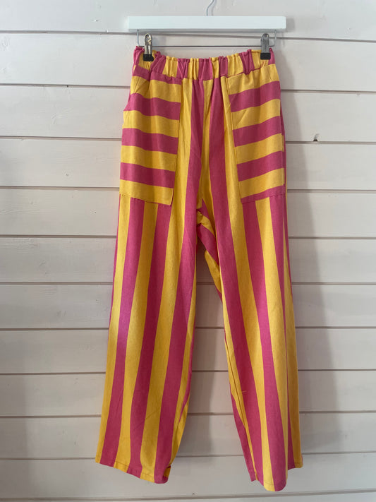 Loose-fitting, vegan-friendly trousers with vivid pink and yellow stripes hang from a white hanger; their elasticated waists and airy fabric guarantee comfort and style against the backdrop of a white wooden wall.