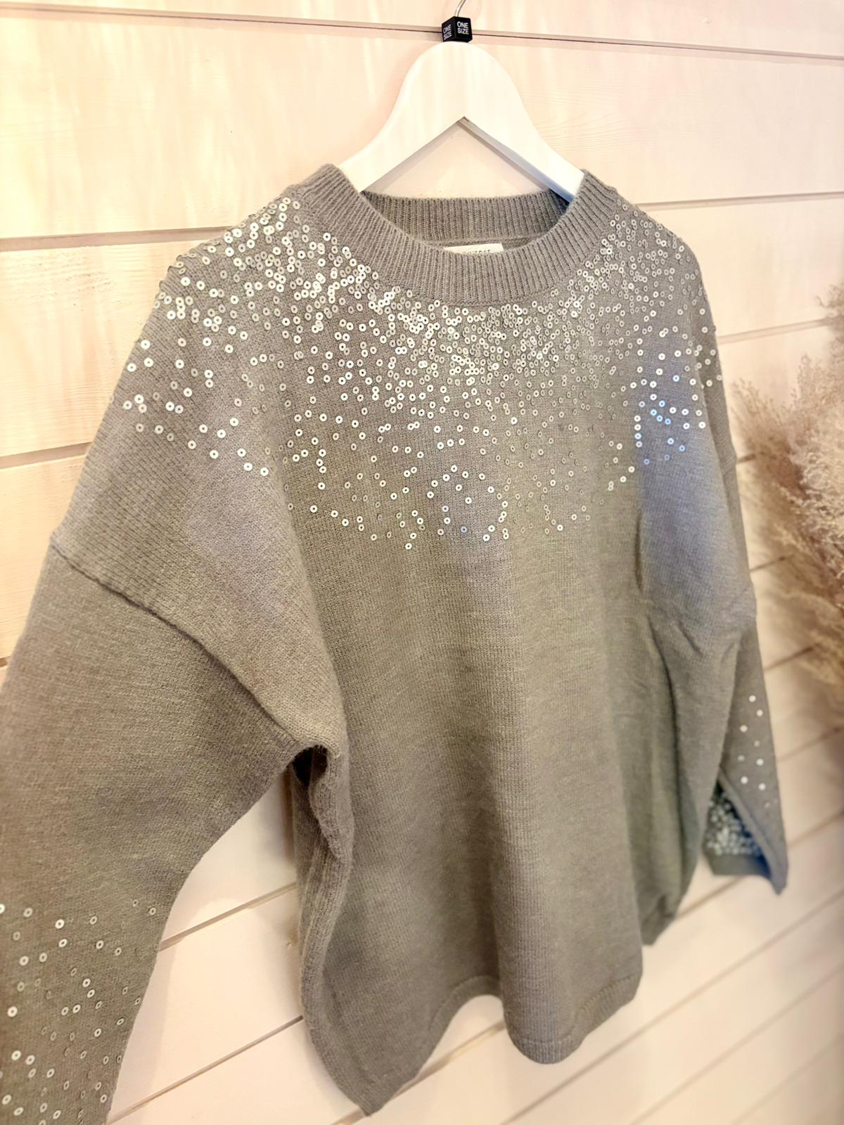 Grey Sequin Jumper featuring silver sequins adorning the shoulders and upper sleeves, ideal for layering. Shown on a white hanger against a light wooden wall.