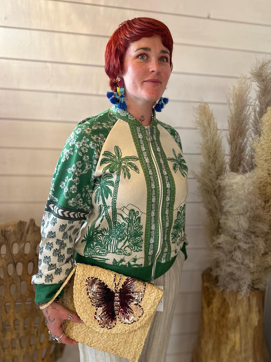 A person with short red hair wears a green and cream patterned jacket, striped trousers, large blue earrings, and holds a straw clutch with a butterfly design, standing by pampas grass.
