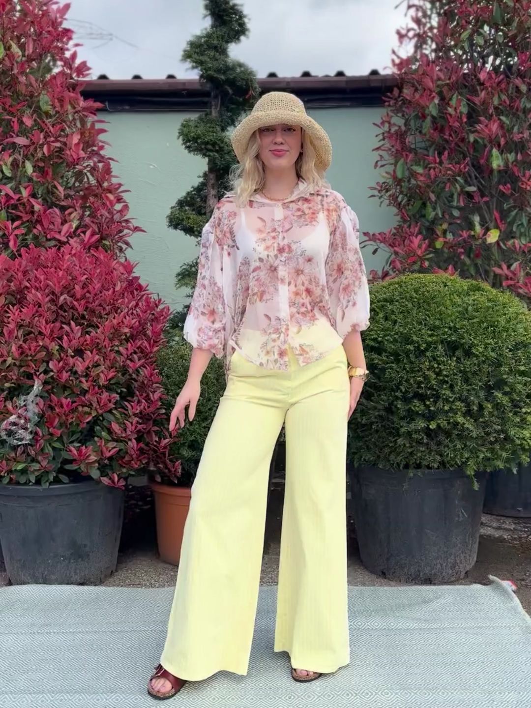 A person stands outdoors on a rug, wearing a straw hat, floral blouse, pinstripe wide-leg jeans and sandals, surrounded by large potted shrubs and red-leaved plants.