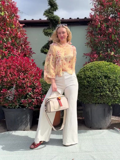 A woman stands outdoors on a rug between topiary bushes, wearing a floral blouse, pinstripe wide-leg jeans, sandals, and carrying a beige handbag.