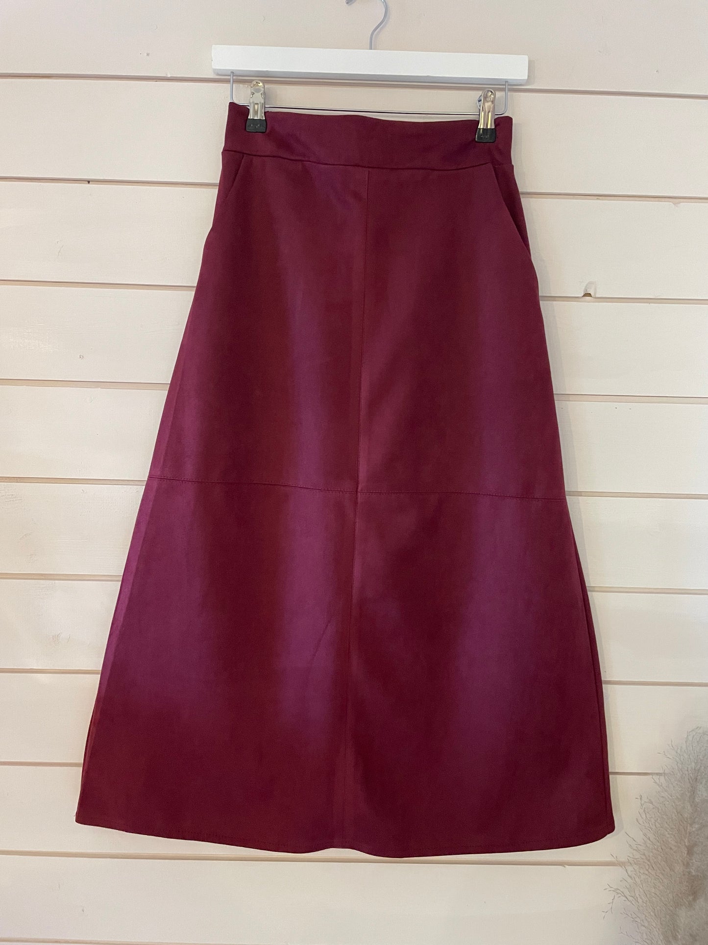 A vegan-friendly, maroon A-line skirt is displayed on a white hanger against a light wooden wall.