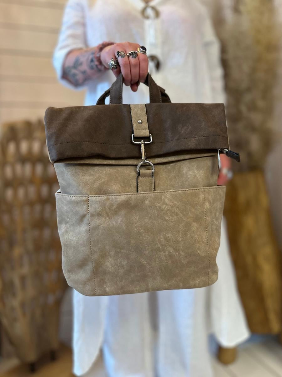 A person in a white outfit holds a beige and brown fold-over canvas rucksack with front pockets and metal fittings, displaying it towards the camera.