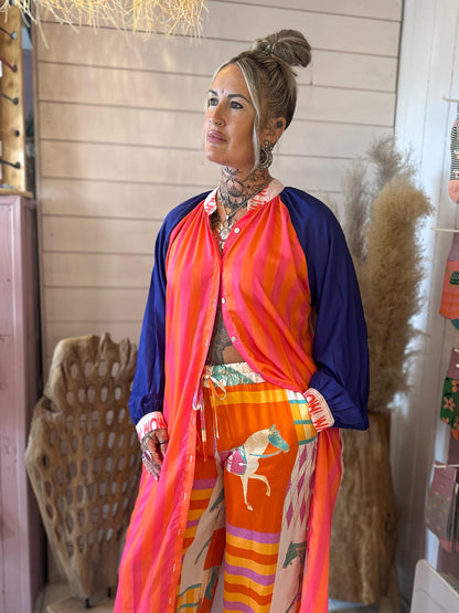 A woman with tattoos is wearing a bright, colourful outfit featuring a striped pink and blue top and orange trousers with horse prints, standing indoors near decorative items.