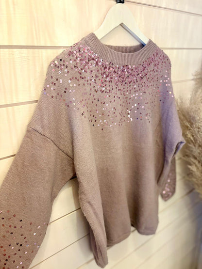 Pale pink jumper with long sleeves, adorned with scattered pink and white sequins around the neckline and shoulders, crafted from vegan materials. Displayed on a white hanger against a wooden wall.