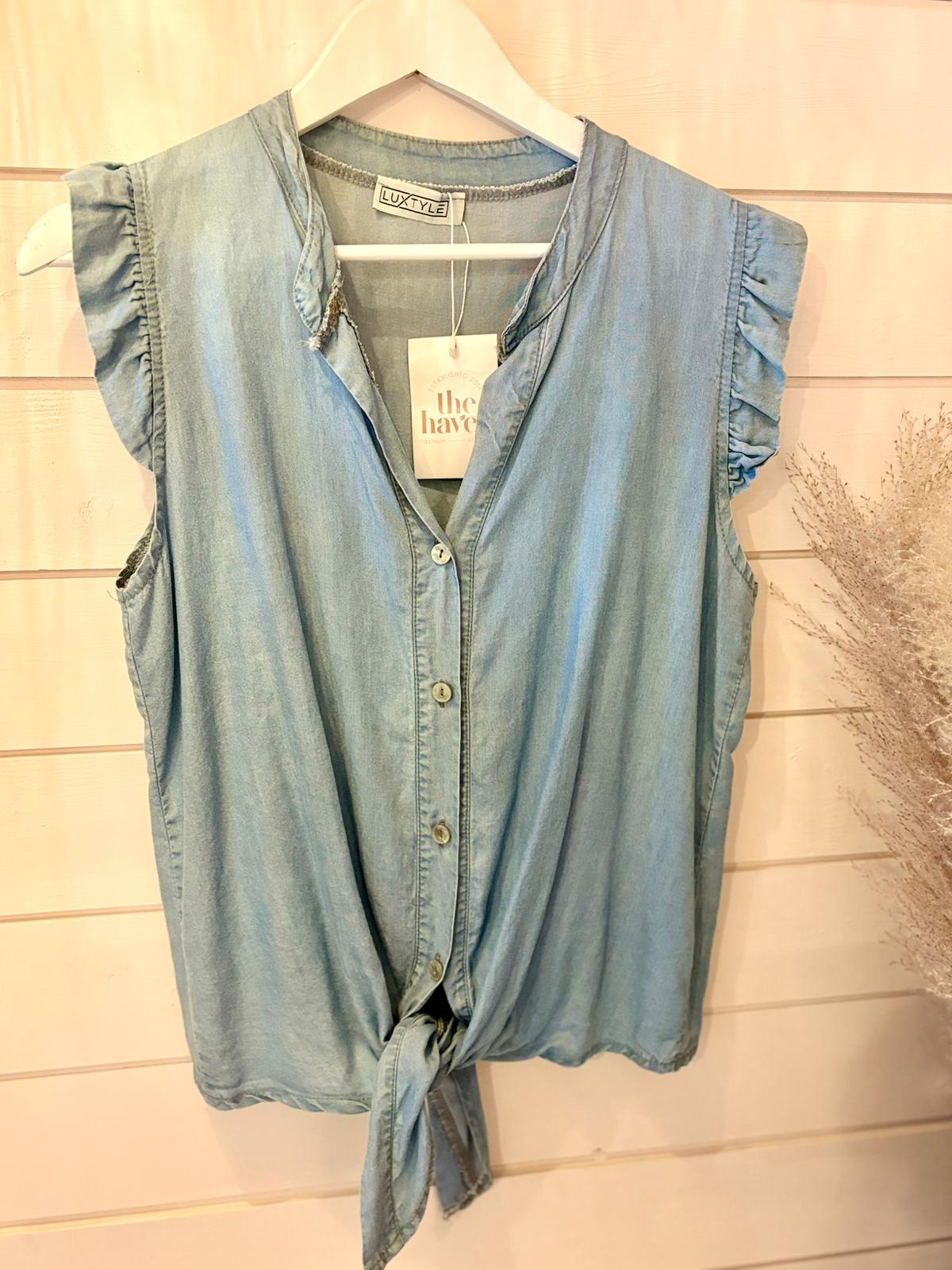 Sleeveless light blue vegan denim blouse featuring ruffled shoulders and a front tie, shown on a hanger against a white wooden wall—a sustainable fashion option.