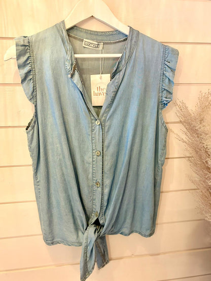 Sleeveless light blue vegan denim blouse featuring ruffled shoulders and a front tie, shown on a hanger against a white wooden wall—a sustainable fashion option.