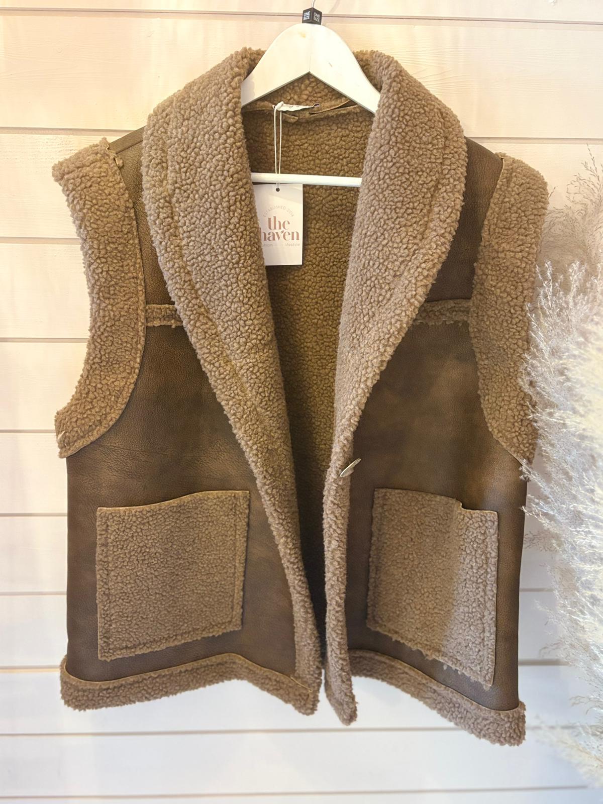 A sleeveless brown faux wool waistcoat with large patch pockets, shown on a white hanger against a light wooden wall.