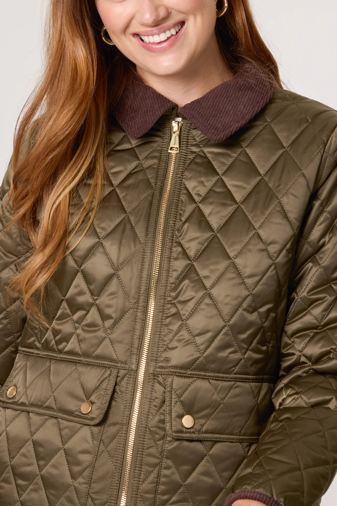 A woman wears a vegan khaki coat with thermal insulation, showcasing an olive green quilted design, brown collar, gold zip fastening, and flap pockets.