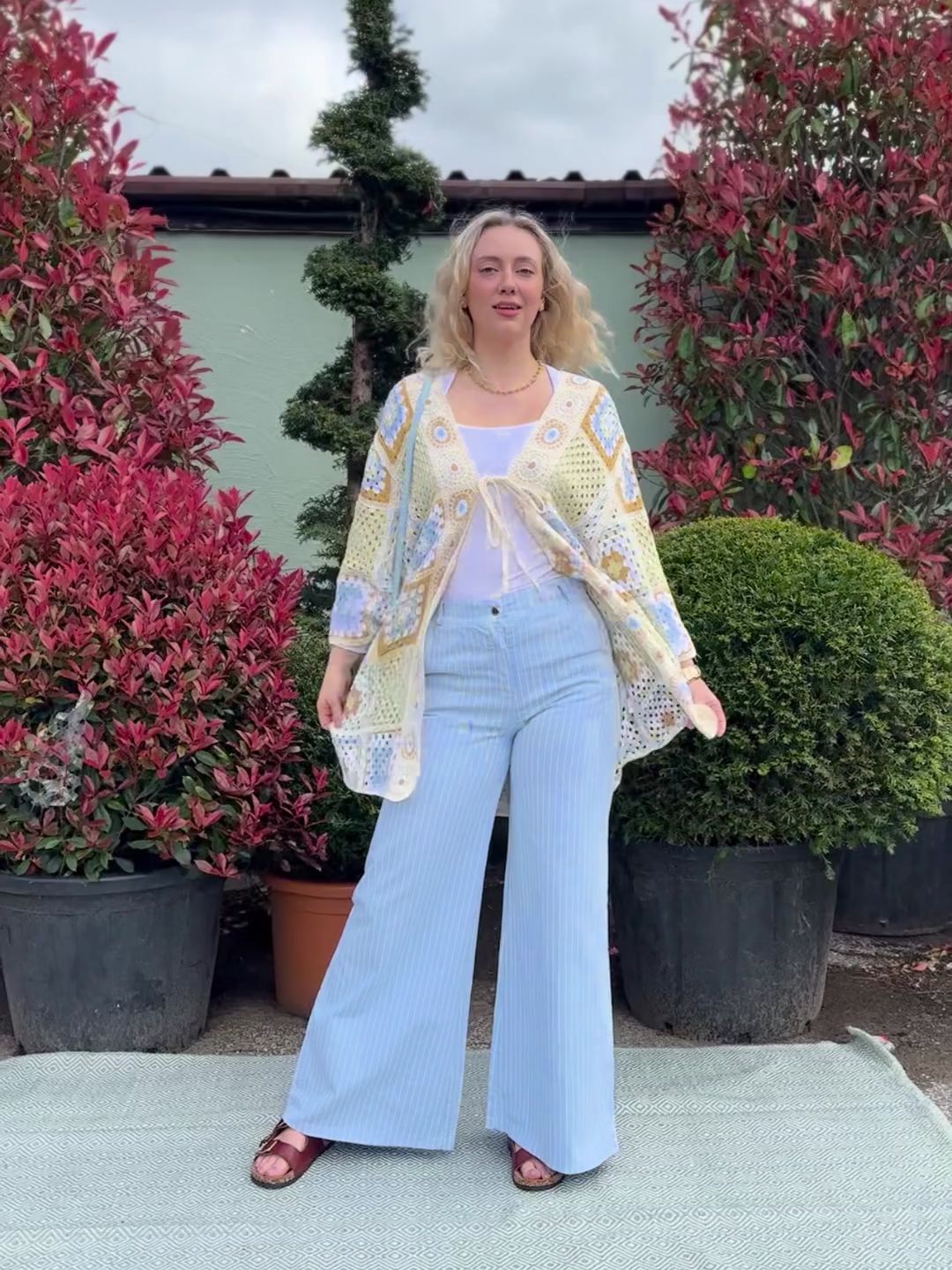 Woman standing outdoors on a rug, wearing light blue pinstripe wide-leg jeans, a white top, and a patterned cardigan, with potted leafy plants in the background.