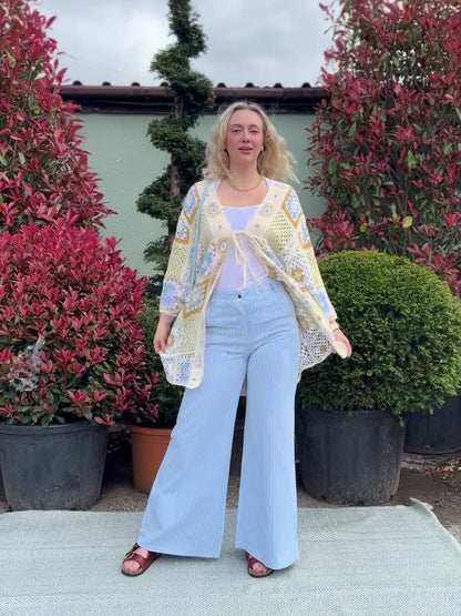 Woman standing outdoors on a rug, wearing light blue pinstripe wide-leg jeans, a white top, and a patterned cardigan, with potted leafy plants in the background.