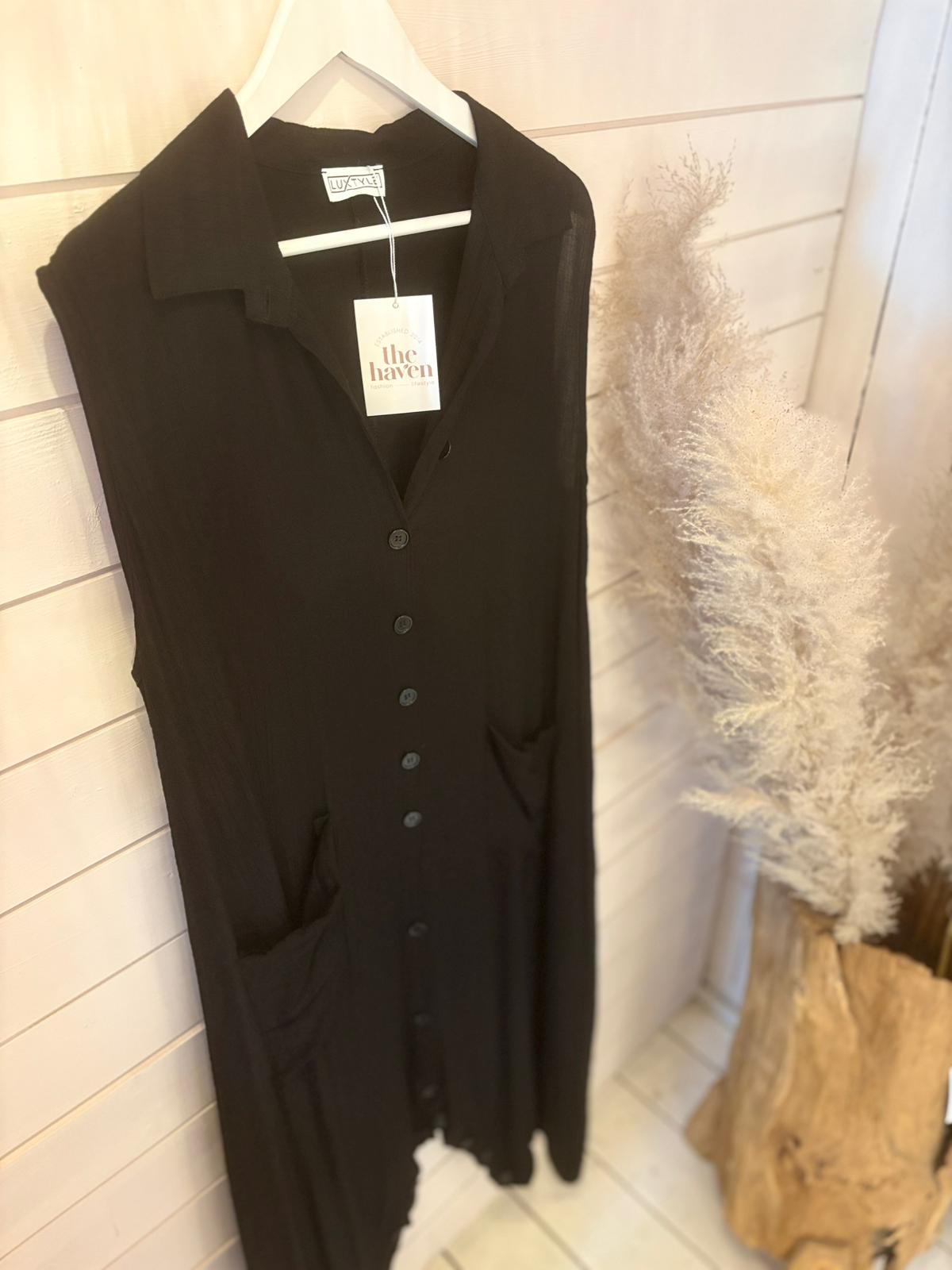 Sleeveless black jumpsuit dress with pockets hangs on a white hanger against a light wooden wall, next to a vase of pampas grass—a versatile jumpsuit ideal for any occasion.