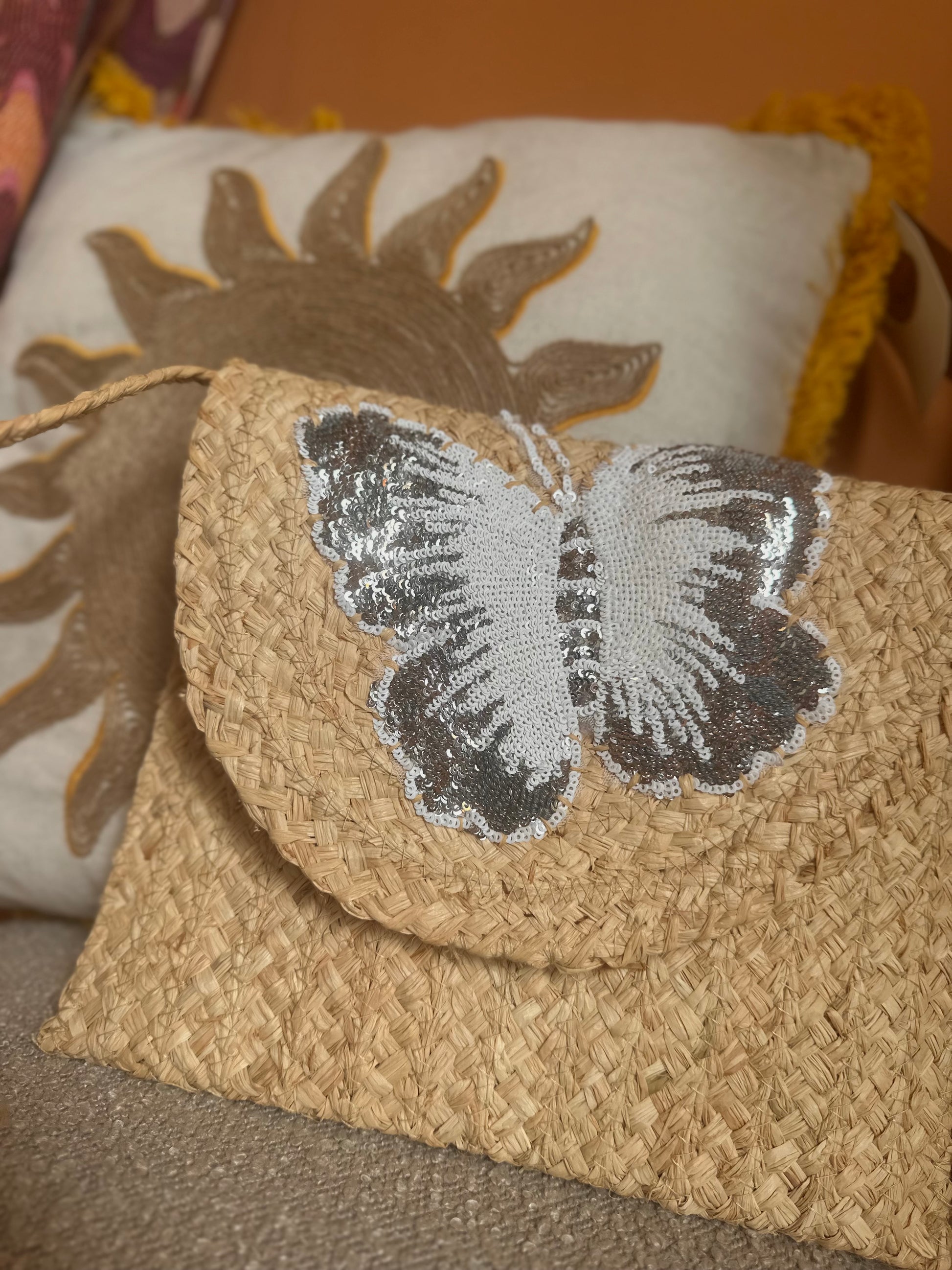 A woven straw bag with a silver and white sequin butterfly on the flap, this stylish handbag sits in front of a decorative cushion featuring a sun design.