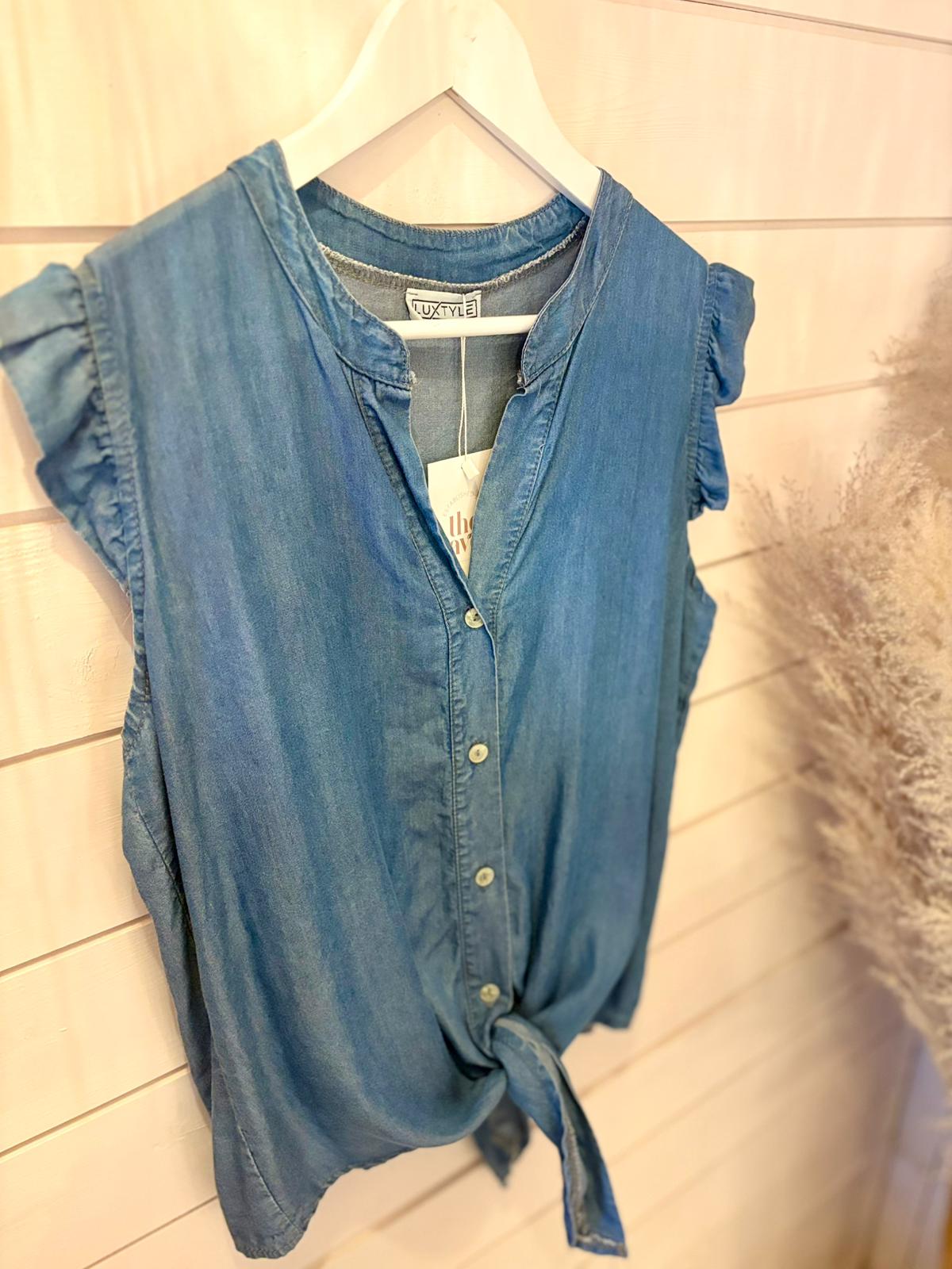 Sleeveless blue chambray button-through top with ruffled shoulders and a front tie, made from eco-friendly vegan materials, hanging on a white hanger against a white panelled wall.