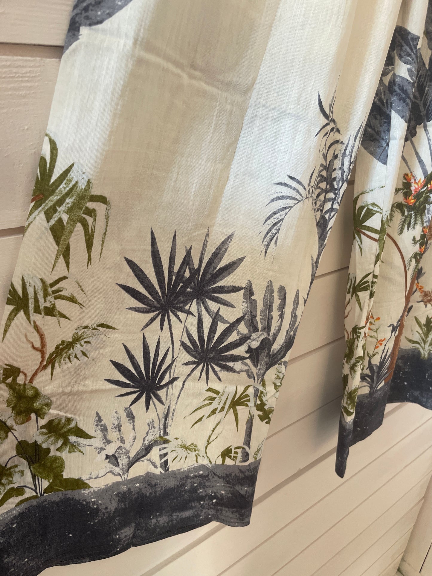 Beige fabric with a printed botanical pattern in green, black, and red hangs against a white wooden wall—ideal for styling with blue jungle trousers or other lightweight trousers with an elasticated waistband.