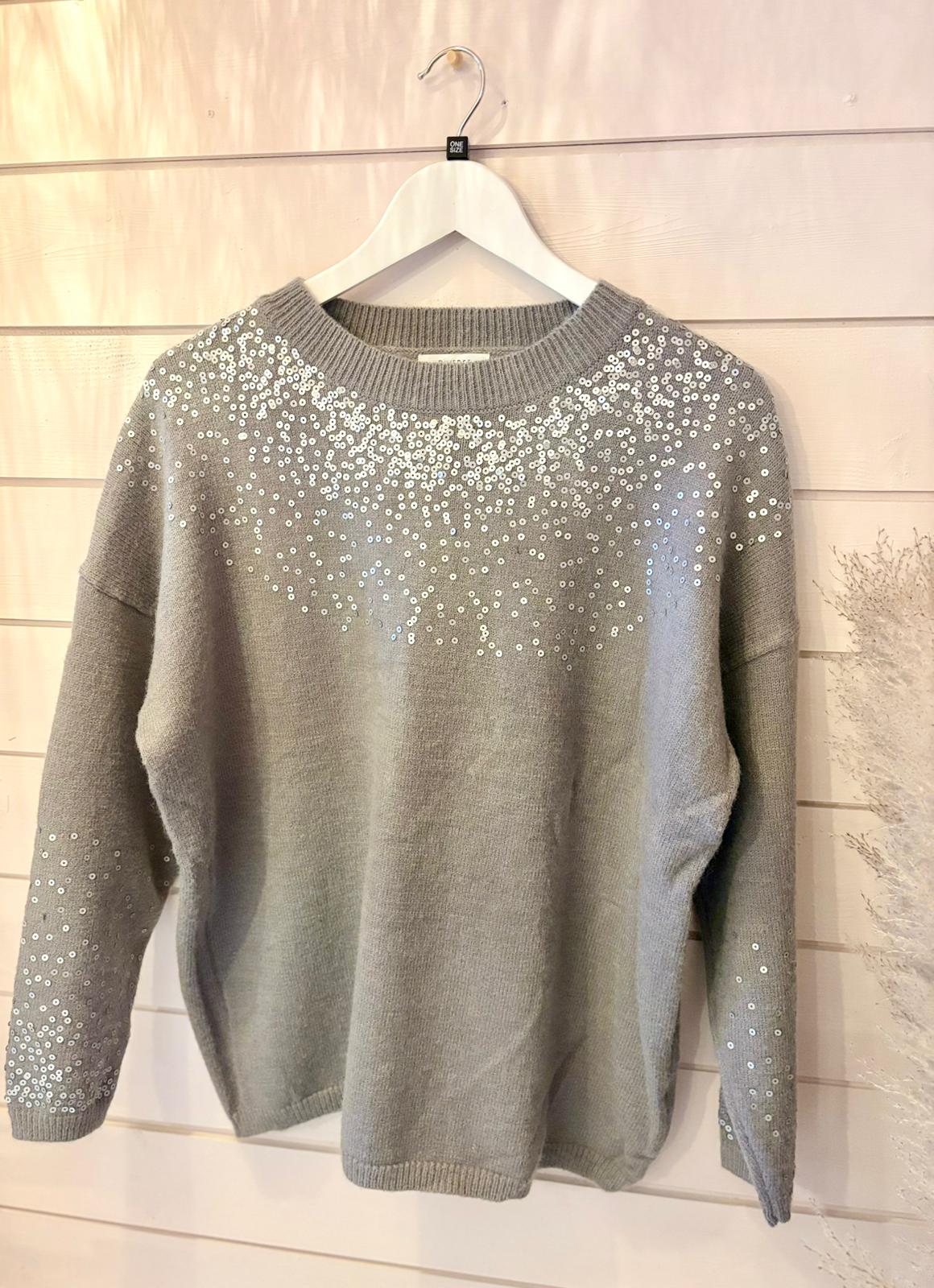 A grey sequin jumper, ideal for layered outfits, hangs on a white hanger against a light wood-panelled wall. This stylish one-size piece is adorned with white sequin accents for extra sparkle.