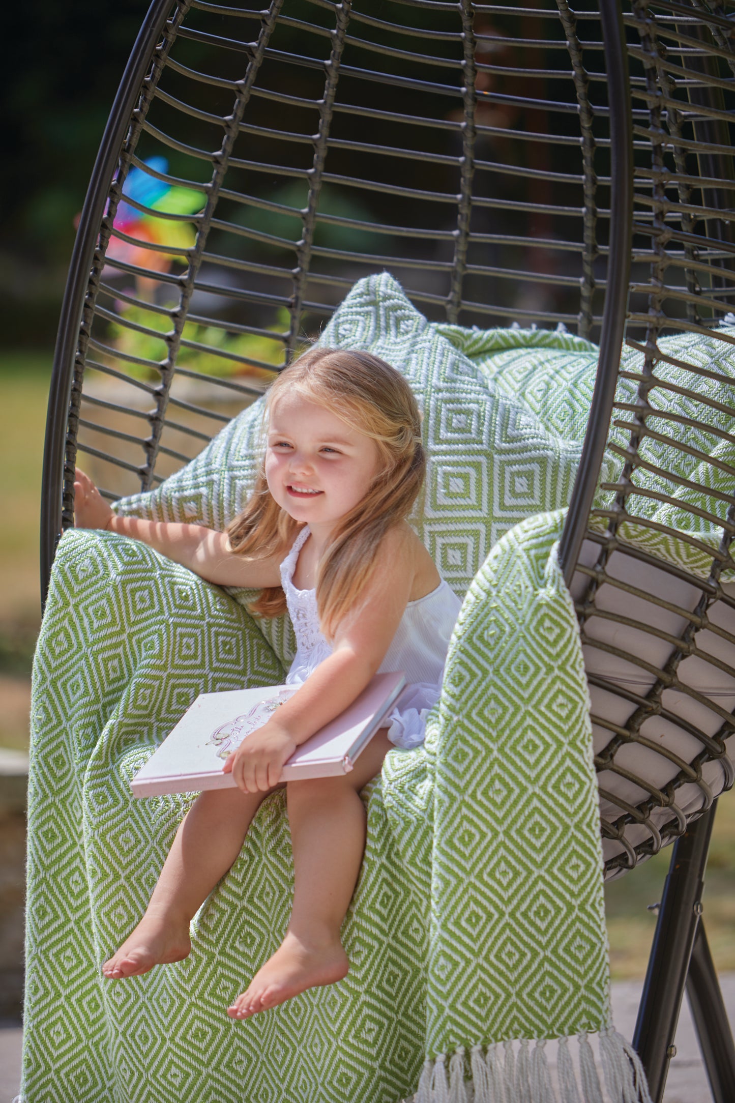 A young girl sits in a hanging chair with a green patterned sustainable throw, made from recycled plastic bottles, holding an open book and smiling outside.