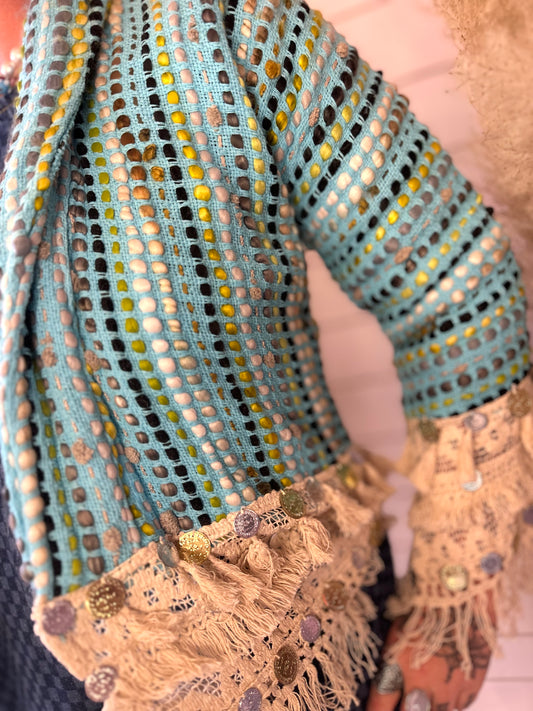 A person is wearing a blue open-knit jumper featuring TBC-inspired cream-coloured, fringed lace and button embellishments on the cuffs and hem.