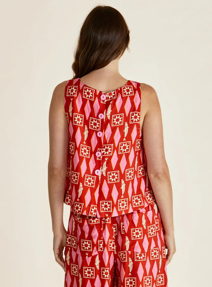 A person with long brown hair is seen from behind wearing a Copacabana Shell Top—a sleeveless red and pink geometric print made from vegan materials—paired with matching shorts.