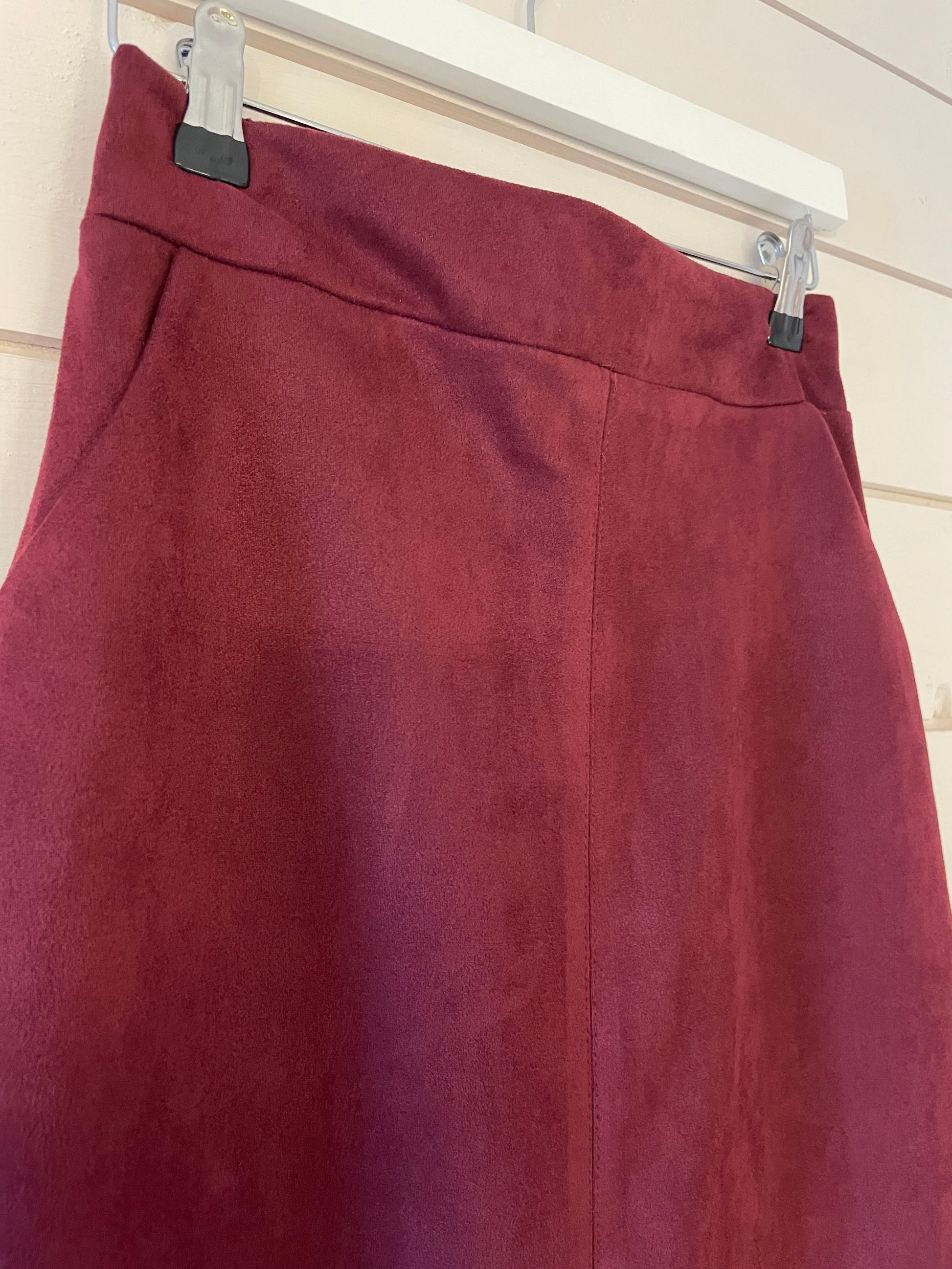 A close-up of a vegan-friendly burgundy skirt hanging on a clear plastic hanger against a light wooden wall.