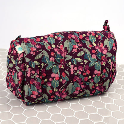 The Winter Berry Quilted Make-up Bag boasts a maroon background with pink and green floral patterns, ideal for keeping your beauty essentials on a chic white hexagonal tiled surface.