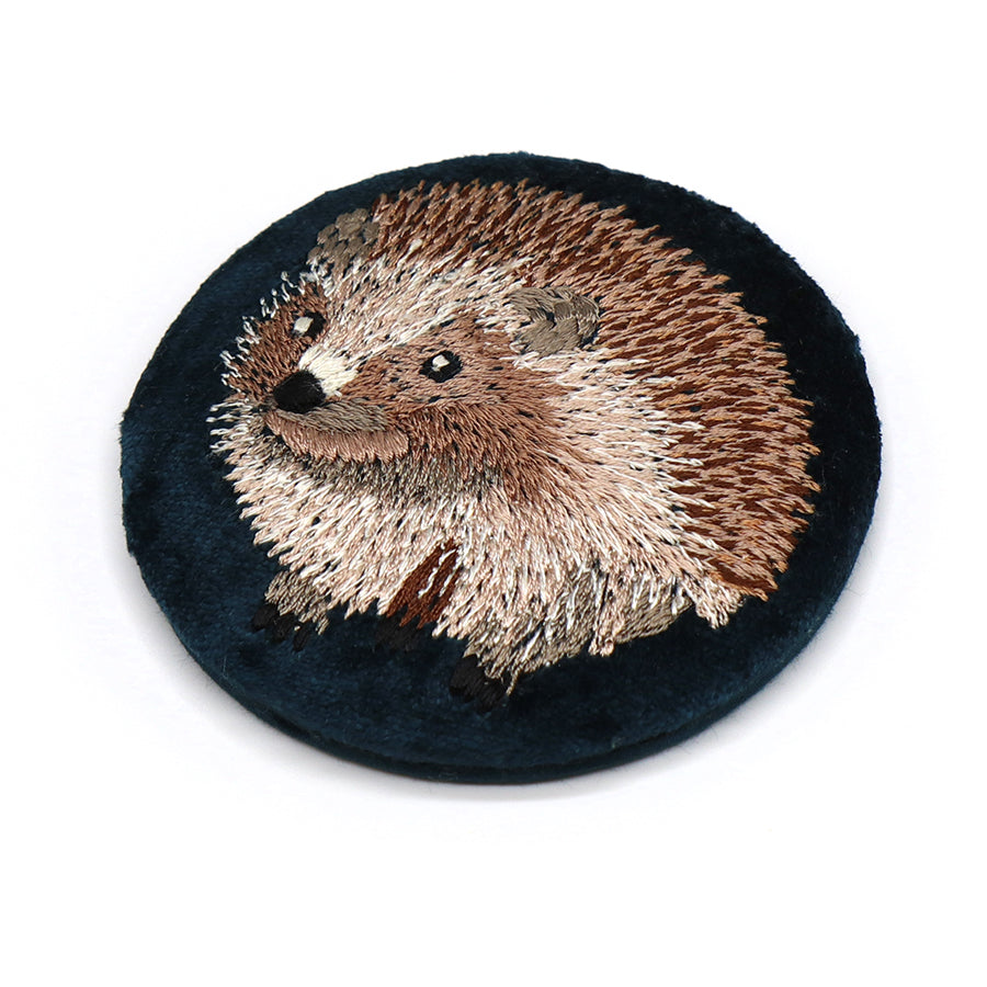 A round black fabric patch displaying an embroidered hedgehog in tones of brown, white, and black—ideal for adding charm to your travel mirror or velvet compact mirror.