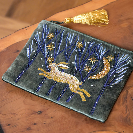A luxurious accessory, this green velvet zip purse boasts an embroidered design of a running hare, trees, stars, a crescent moon, and a gold tassel zip pull, set atop a wooden surface.