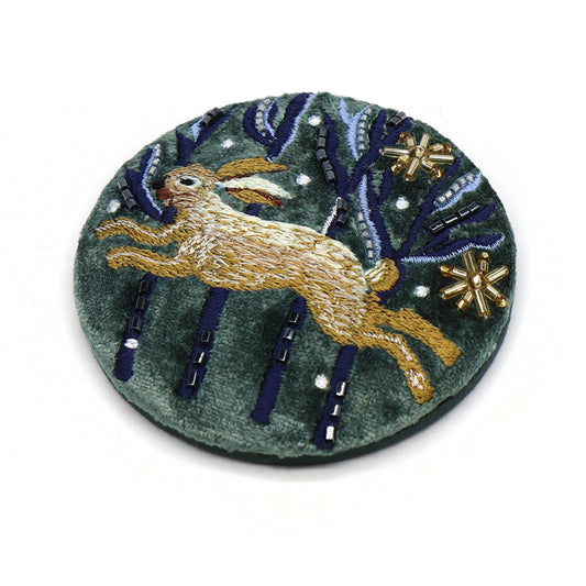 A circular embroidered accessory patch depicting a brown rabbit leaping amid blue and green foliage, embellished with gold and white bead details.