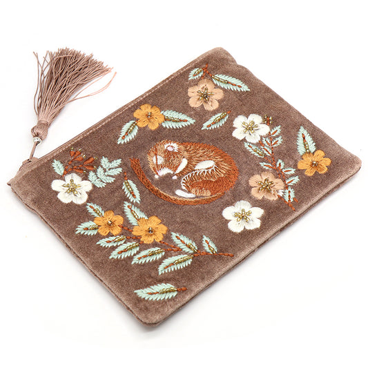 A brown velvet zipped purse with an embroidered motif of a curled-up fox surrounded by yellow and white flowers, finished with a gold tassel on the zip.