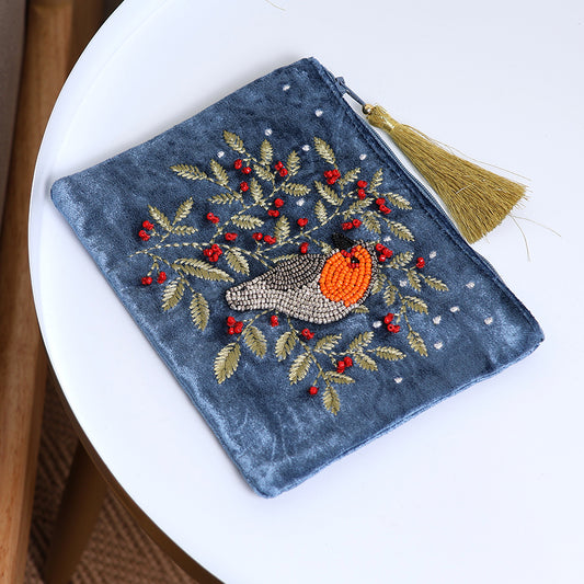 A blue velvet zip purse featuring an embroidered bird and foliage, red berries, small white dots, and a gold tassel zip, resting on a white round table.