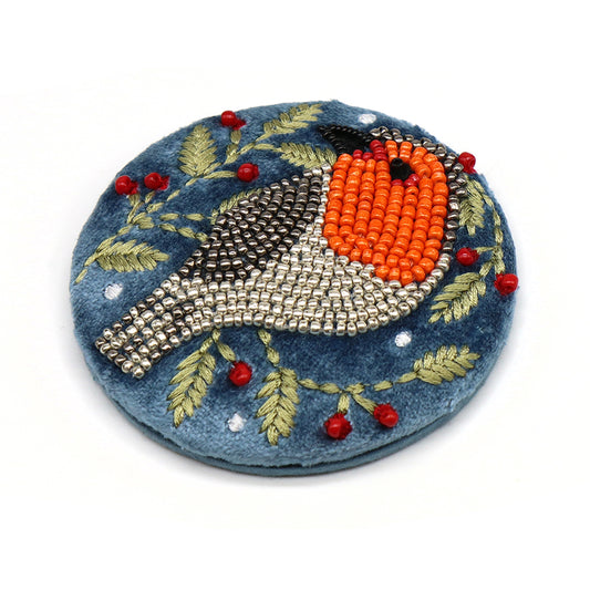 A round embroidered brooch accessory featuring a bird motif crafted with beads and thread on blue fabric, embellished with leaves and small red berries.