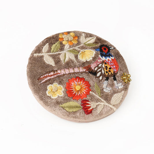 A round, brown felt brooch adorned with colourful embroidered flowers and a bird, reminiscent of patterns seen on an embroidered mirror, displayed against a white background.