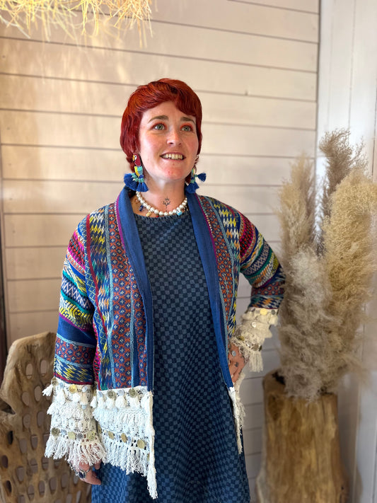A woman with short red hair is wearing a colourful, patterned jacket over a blue dress, standing indoors next to pampas grass and wood-panelled walls.