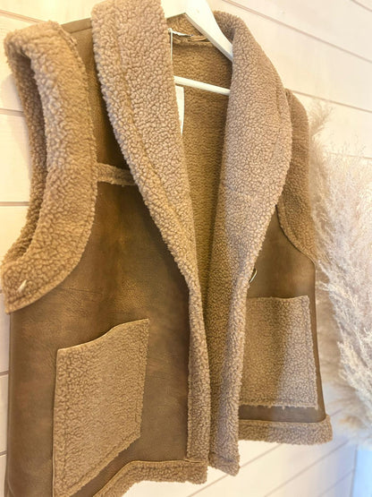 A brown faux wool waistcoat with large front pockets hangs on a white hanger against a light wooden wall.