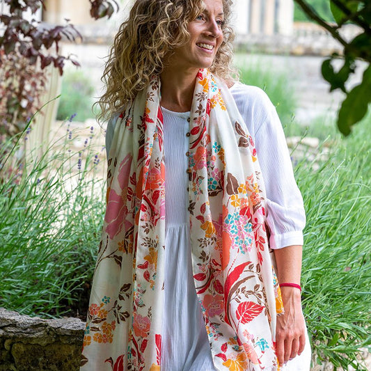 Woman with curly hair wearing a white dress and a Natural Summer Scarf crafted from vegan materials, standing outdoors beside greenery and stone.
