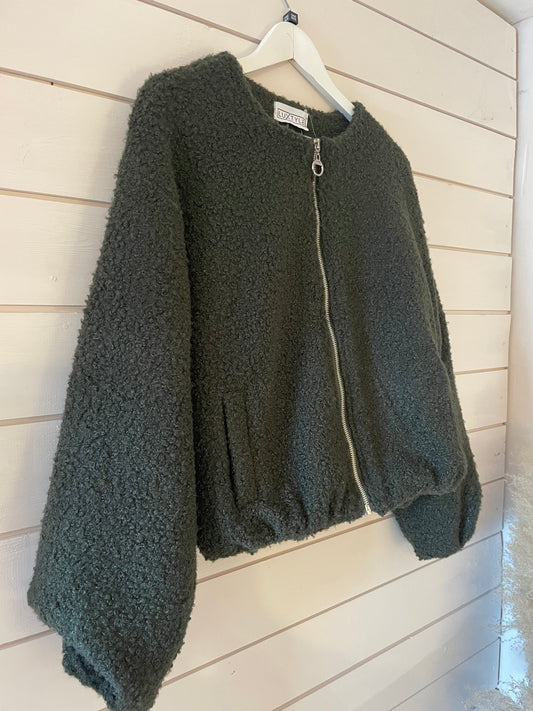 A vegan-friendly forest green bomber jacket with wide sleeves and textured fabric hangs on a white hanger against a white wooden wall.