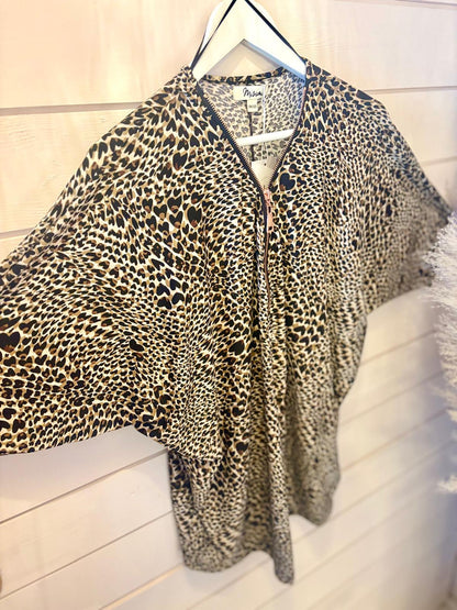 A loose-fitting, short-sleeved animal print blouse with a zip front is shown on a hanger against a white wooden wall.