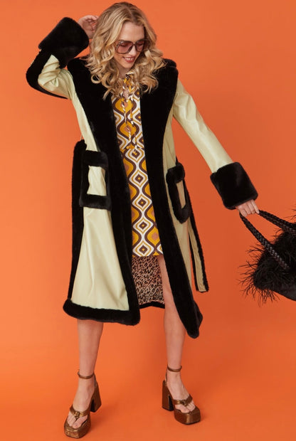 A woman in a long, light-coloured coat with black trim and leopard print lining, a retro-patterned dress, platform heels, and sunglasses stands against an orange background holding a black handbag.